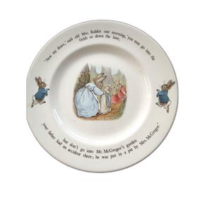 May include: A white ceramic plate featuring illustrations of Peter Rabbit characters. The center shows a rabbit in a blue coat with another rabbit. Two smaller rabbits in blue coats are on the sides. Text reads: Now my dears, said old Mrs. Rabbit one morning, you may go into the fields or down the lane, but don't go into Mr. McGregor's garden your father had an accident there; he was put in a pie by Mrs. McGregor.