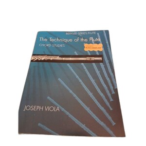 May include: A music instruction book titled The Technique of the Flute: Chord Studies by Joseph Viola. The cover is dark blue with a horizontal photograph of a silver flute and several light blue diagonal outlines of flutes across the front.