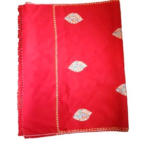 May include: A vibrant red textile with intricate embroidery. The fabric features a repeating pattern of stylized leaf-like designs in shades of blue, yellow, and white. The edges are finished with a decorative fringe and a detailed border.