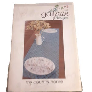May include: A rectangular product packaging for gailpan designs titled my country home. The cover features a photograph of a blue textured table runner with white oval embroidered patches depicting a house, trees, and a heart, plus a white pitcher.
