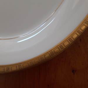 May include: A white ceramic plate with a gold rim and a geometric pattern. The plate is slightly curved and has a glossy finish.