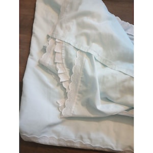 May include: A light blue bed sheet set with white lace trim. The set includes a flat sheet and a pillowcase, both with delicate scalloped lace edges. The sheets are folded, showing the details of the fabric and trim.