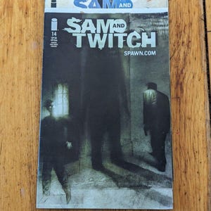 May include: A Sam and Twitch issue 14 comic book with a dark, moody cover featuring a large central shadow, a figure standing on the left, and a figure walking away on the right. The title Sam and Twitch and the website SPAWN.COM are printed.