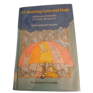 May include: A children's book titled "It's Raining Cats and Dogs" with illustrations by True Kelley. The cover features a colorful umbrella sheltering two children from falling cats and dogs. The book's title and author's name are visible.