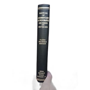 May include: A black book titled "Manual of Sedimentary Petrography" with gold lettering and accents. The book is a classic facsimile edition and part of the SEPM reprint series, number 13. The book is held by a hand.