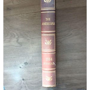 May include: A vintage hardcover book titled "THE AMERICANA 1964 ANNUAL." The book has a dark burgundy leather cover with gold lettering and decorative accents. The spine is visible, showing the title and year.