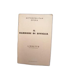 May include: A vintage booklet for "Il Barbiere di Siviglia" from the Metropolitan Opera. The cover is a light beige color with black text. The title and "Libretto" are prominently displayed.