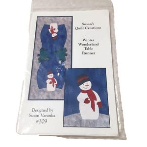 May include: A package for a Winter Wonderland Table Runner designed by Susan Varanka. The package features a blue table runner design with snowman and tree illustrations. The text "Susan's Quilt Creations" is visible.