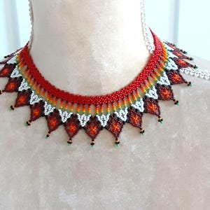 May include: A red, white, and brown beaded necklace with a geometric pattern. The necklace is made of small seed beads and has a scalloped edge.