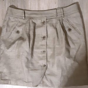 May include: A beige-colored skirt with a button-down front closure. The skirt features two front pockets with button details and a waistband with belt loops. The fabric has a subtle textured pattern, and the skirt appears to be knee-length.