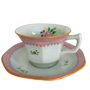 May include: A vintage teacup and saucer set. The cup and saucer are white with a light blue tint, featuring a pink border with a geometric design and orange trim. Floral details adorn the cup and saucer.