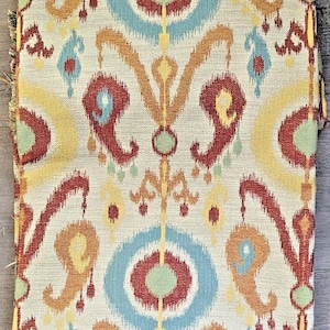 May include: A rectangular piece of fabric featuring a repeating ikat pattern in shades of rust red, light blue, mustard yellow, and pale green on a cream-colored background. The textile displays symmetrical, stylized paisley and circular motifs.