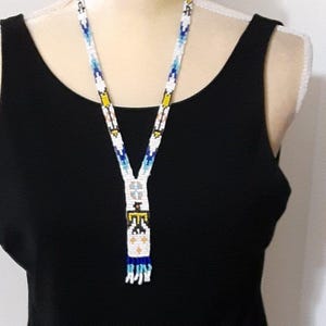 May include: A long beaded necklace featuring a white base with geometric patterns in blue, yellow, and black. The design includes a central vertical section with a stylized bird motif and a fringe of blue and white beads at the bottom end.