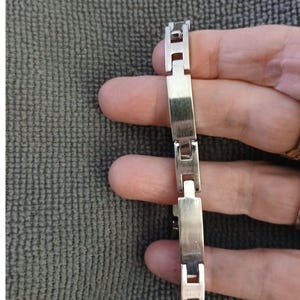 May include: A silver-toned bracelet with rectangular links and a clasp. The bracelet is made of metal and has a polished finish. The links are connected by smaller, square-shaped pieces.