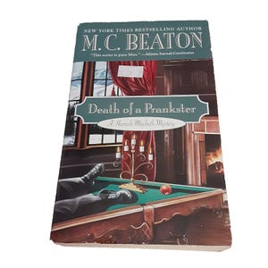 May include: Book cover for "Death of a Prankster" by M.C. Beaton, a Hamish Macbeth Mystery. The cover features a painting of a man lying on a pool table in a room with a fireplace and a window. The book is a New York Times Bestselling Author.