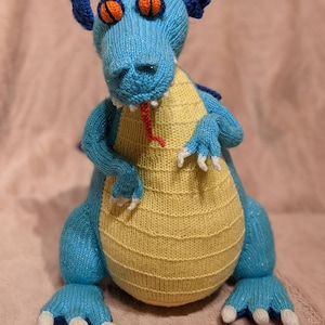 May include: A blue and yellow knitted dragon toy with orange eyes and a red tongue. The dragon is sitting on a pink fuzzy surface.