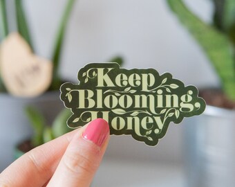 Puede incluir: Una pegatina verde con el texto "Keep Blooming, Honey" en una fuente de color amarillo claro. La pegatina tiene un diseño floral alrededor del texto.