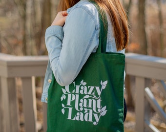 Puede incluir: Una bolsa de tela verde con texto blanco que dice "Crazy Plant Lady". La bolsa tiene una correa blanca y la está sosteniendo una persona.