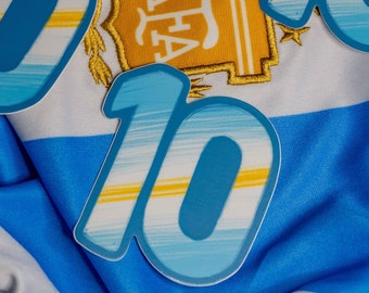 Puede incluir: Un parche azul y amarillo con el número 10 sobre una tela a rayas blanca y azul claro. El parche tiene un efecto descolorido, como de pincelada.