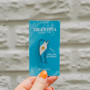 Può includere: Uno spillo in smalto blu e bianco con un contorno dell'Argentina con i colori della bandiera del paese e un sole al centro. Lo spillo dice "Argentina Enamel Pin" e "Que Viva La Patria" in testo bianco.