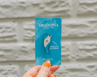 Puede incluir: Un pin de esmalte azul y blanco con un contorno de Argentina con los colores de la bandera del país y un sol en el centro. El pin dice "Argentina Enamel Pin" y "Que Viva La Patria" en texto blanco.