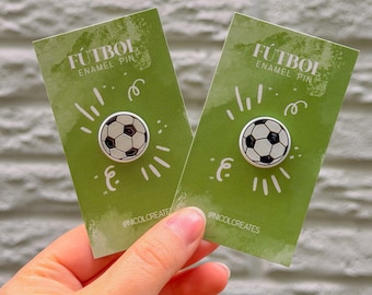 Puede incluir: Dos pines de esmalte con un diseño de balón de fútbol. Cada pin está en una tarjeta verde con la palabra "FÚTBOL" y el texto "ENAMEL PIN". Fondo de pared de ladrillo blanco.