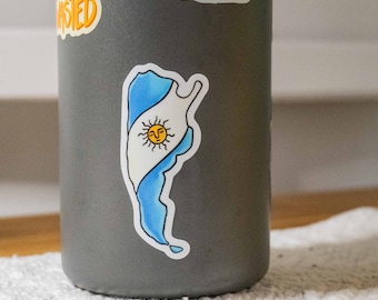 Puede incluir: Una botella de agua gris con una pegatina del contorno de Argentina con la bandera argentina en su interior. La bandera es azul y blanca con un sol amarillo en el centro.
