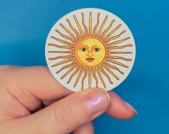 Puede incluir: Una pegatina redonda con un sol estilizado con una cara. El sol tiene una cara y rayos amarillos, sobre un fondo blanco. La pegatina se sostiene sobre un fondo azul.
