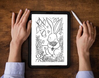 Puede incluir: Dibujo lineal en blanco y negro de un conejito sosteniendo flores, rodeado de flores, corazones y una mariposa. La obra de arte se muestra en una tableta, sostenida por manos, con un lápiz óptico. La tableta está sobre una superficie de madera.