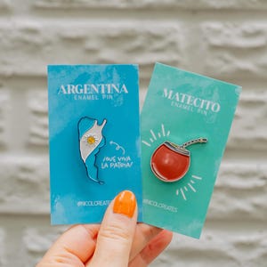 Może przedstawiać: Dwa szpilki emaliowane na niebieskich i turkusowych kartkach. Lewa szpilka przedstawia biały zarys Argentyny z flagą kraju w środku. Tekst "Argentina Enamel Pin" i "¡Que viva la patria!" znajduje się na kartce. Prawa szpilka przedstawia czerwoną i białą filiżankę do mate z rurką. Tekst "Matecito Enamel Pin" znajduje się na kartce.