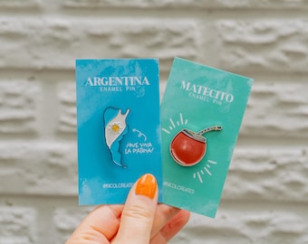 Puede incluir: Dos pines de esmalte en tarjetas azules y turquesa. El pin de la izquierda presenta un contorno blanco de Argentina con la bandera del país en su interior. El texto "Argentina Enamel Pin" y "¡Que viva la patria!" está en la tarjeta. El pin de la derecha presenta una taza de mate roja y blanca con una pajita. El texto "Matecito Enamel Pin" está en la tarjeta.