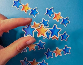 Puede incluir: Pegatinas en forma de estrella en azul, naranja y amarillo. Las pegatinas están dispuestas sobre un fondo azul, algunas sujetas por una mano. Las estrellas tienen un borde blanco.