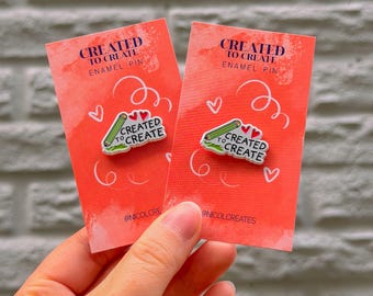 Puede incluir: Dos tarjetas rojas con texto blanco que dice "CREATED TO CREATE ENAMEL PIN". Cada tarjeta tiene un pin con un lápiz verde y las palabras "CREATED TO CREATE". Corazones blancos y espirales decoran las tarjetas.