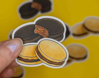 Puede incluir: Hoja de pegatinas con pegatinas de alfajores. Las pegatinas representan galletas redondas con una cobertura de chocolate negro y un relleno de dulce de leche. Algunas pegatinas están enteras, otras muestran una sección transversal. El fondo es amarillo.