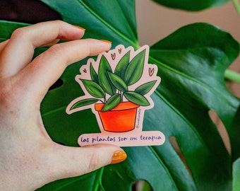 Puede incluir: Una pegatina rosa con una ilustración dibujada a mano de una planta en maceta con hojas verdes y una maceta de terracota. La pegatina tiene el texto "las plantas son mi terapia" en escritura cursiva negra.
