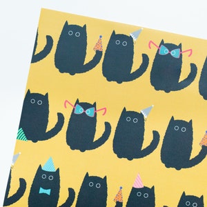 May include: A yellow wrapping paper with a repeating pattern of black cats wearing party hats and glasses. Some cats are wearing bow ties.