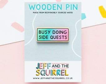 Op de afbeelding: Een houten pin met een blauwe en roze gradiënt achtergrond en gouden rand. De pin zegt "Busy Doing Side Quests". De pin is gemaakt door Jeff and the Squirrel.
