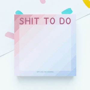 Puede incluir: Un bloc de notas cuadrado con el texto "SHIT TO DO" en color malva. El bloc de notas tiene un diseño degradado con rayas diagonales rosas, azules y blancas. Las palabras "JEFF AND THE SQUIRREL" están impresas en la parte inferior.