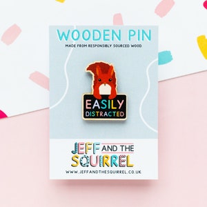 May include: Wooden pin featuring a cartoon squirrel holding a sign that reads "Easily Distracted". The pin is made from responsibly sourced wood and is a product of Jeff and the Squirrel.