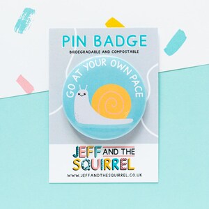 May include: A pin badge with a cartoon snail design. The badge is light blue with a yellow snail shell and the text "GO AT YOUR OWN PACE". The badge is on a white card with the text "PIN BADGE" and "JEFF AND THE SQUIRREL".