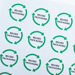 May include: A sheet of green and white circular stickers with the text "REUSED PACKAGING" printed on each sticker. The stickers feature a green circular arrow design.