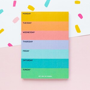 May include: A rectangular weekly planner notepad featuring seven horizontal colour-blocked sections in yellow, orange, pink, light purple, light blue, teal, and green. Each section is labelled with a day of the week and the text JEFF AND THE SQUIRREL.