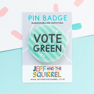 Può includere: Un distintivo rotondo con un design a righe verdi e bianche e le parole "VOTE GREEN" in nero. Il distintivo è su una carta bianca con il testo "PIN BADGE" e "JEFF AND THE SQUIRREL". Lo sfondo è azzurro.