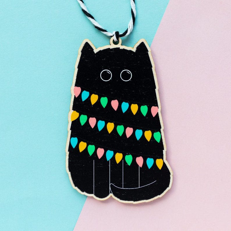 May include: Black wooden cat ornament with colorful string lights wrapped around it. The cat has a white string hanging from the top for hanging.