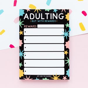May include: A black notepad with the words "ADULTING >BUT WITH REWARDS<" in white. The notepad has a section for rewards and is decorated with colourful stars. The text "JEFF AND THE SQUIRREL" is at the bottom.