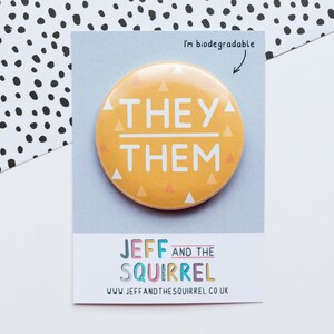 May include: A round, yellow button with the words "THEY/THEM" in white, surrounded by small triangles. The button is on a gray card with the text "JEFF AND THE SQUIRREL" and "I'm biodegradable". The background has a black and white polka dot pattern.