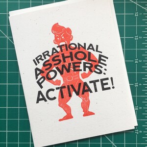 May include: A red and black illustrated greeting card with a cartoon man flexing his muscles and the text "IRRATIONAL ASSHOLE POWERS: ACTIVATE!"