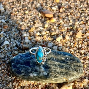 Könnte beinhalten: Ein silberner Ring mit einem ovalen türkisfarbenen Stein und einer Ansammlung kleiner silberner Perlen. Der Ring liegt auf einem dunkelgrauen Stein, vor einem sandigen Hintergrund.