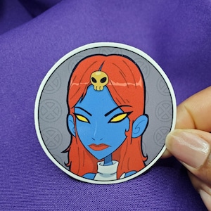 May include: A round sticker featuring a blue-skinned character with red hair and a skull-shaped headpiece. The character has yellow eyes and a stern expression. The sticker is held against a purple fabric background.