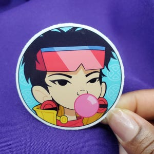 May include: A round sticker featuring a cartoon character with black hair, red visor, and a pink bubble gum bubble. The character is wearing a yellow jacket and red shirt. The sticker has a light blue background with a white border.